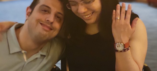 A woman is seated in a wheelchair. She has dark hair and purple glasses, and is wearing a black sleeveless top and purple skort. She is holding her left hand, which displays an engagement ring on her ring finger. Next to her is her fiancé, a man with dark hair and wearing a light blue polo and navy blue jeans. They are smiling.